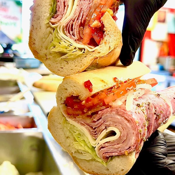Gloved hands hold two halves of a deli sub sandwich filled with meats, cheese, lettuce, and tomatoes.