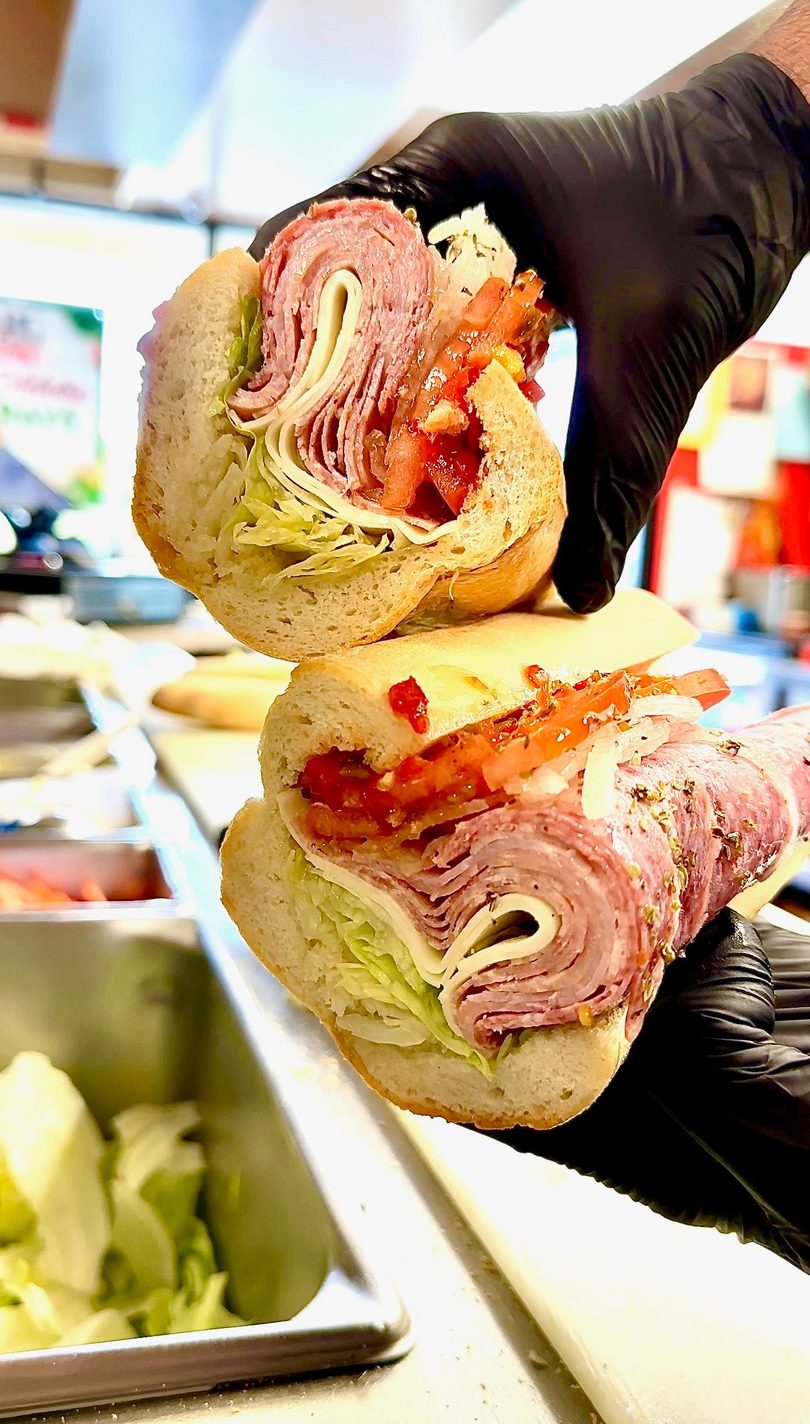 Gloved hands hold two halves of a deli sub sandwich filled with meats, cheese, lettuce, and tomatoes.