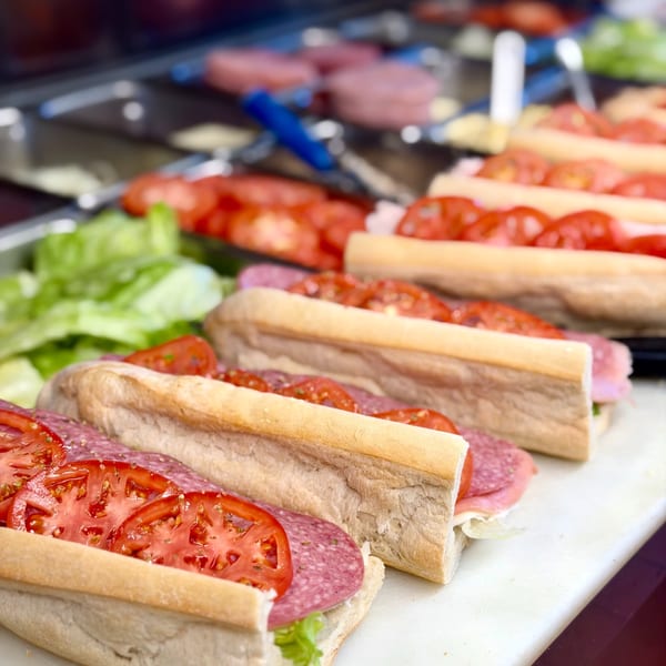 Multiple fresh deli sandwiches with salami, lettuce, and sliced tomatoes on a white cutting board.
