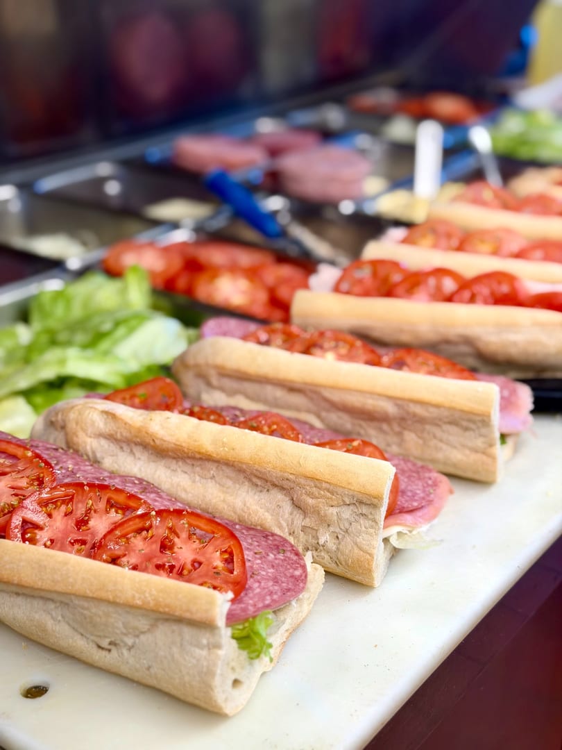 Multiple fresh deli sandwiches with salami, lettuce, and sliced tomatoes on a white cutting board.