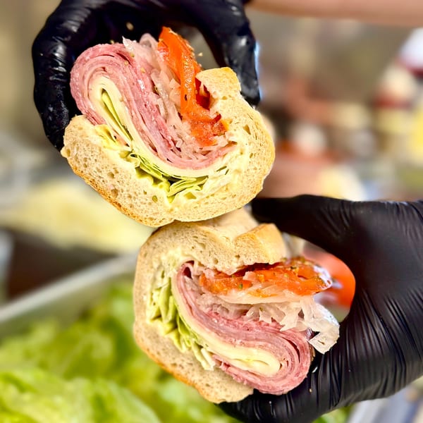 Gloved hands hold a sliced Italian sub sandwich with meats, cheese, lettuce, and toppings.