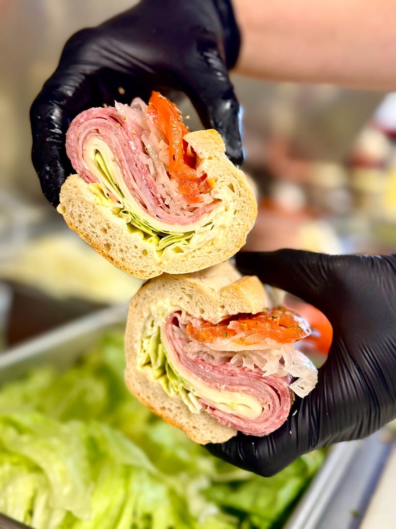 Gloved hands hold a sliced Italian sub sandwich with meats, cheese, lettuce, and toppings.