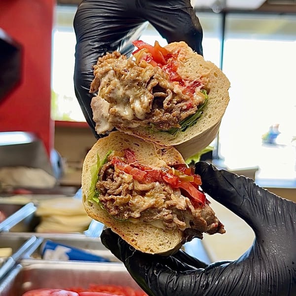 Gloved hands hold a sliced cheesesteak sandwich with melted cheese, steak, lettuce, and tomatoes.