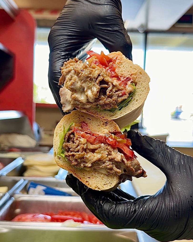 Gloved hands hold a sliced cheesesteak sandwich with melted cheese, steak, lettuce, and tomatoes.