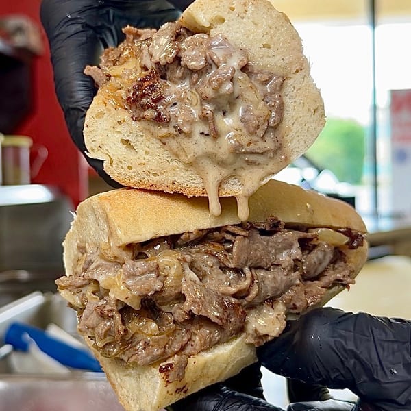 Gloved hands hold a sliced cheesesteak sandwich, revealing melted cheese, steak, and onions.