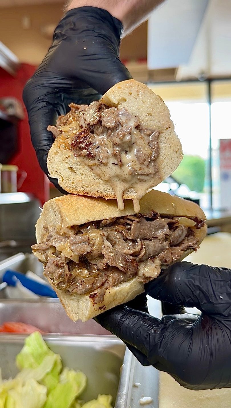 Gloved hands hold a sliced cheesesteak sandwich, revealing melted cheese, steak, and onions.