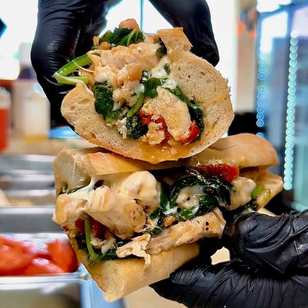 Gloved hands hold a chicken, spinach, and cheese sub sandwich, split in half, in a restaurant setting.
