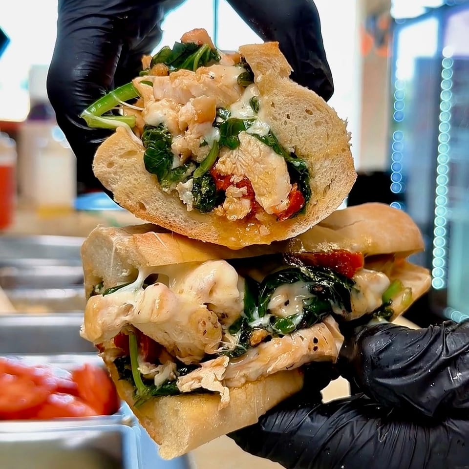 Gloved hands hold a chicken, spinach, and cheese sub sandwich, split in half, in a restaurant setting.