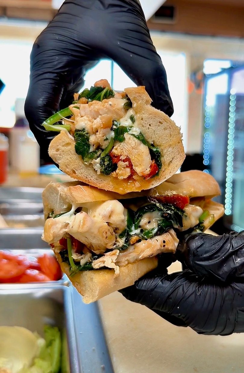 Gloved hands hold a chicken, spinach, and cheese sub sandwich, split in half, in a restaurant setting.
